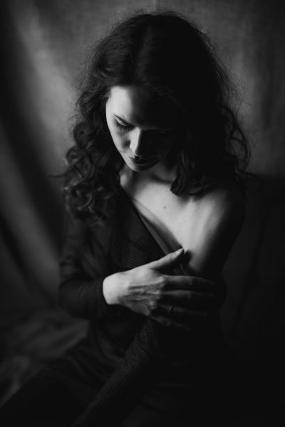 Sinnliches schwarz weiß Foto einer Frau die den Blick nach unten richtet und deren rechte Hand den Saum des über die linke Schulter gerutschten schwarzen Kleides umfasst im Studio in Hannover.